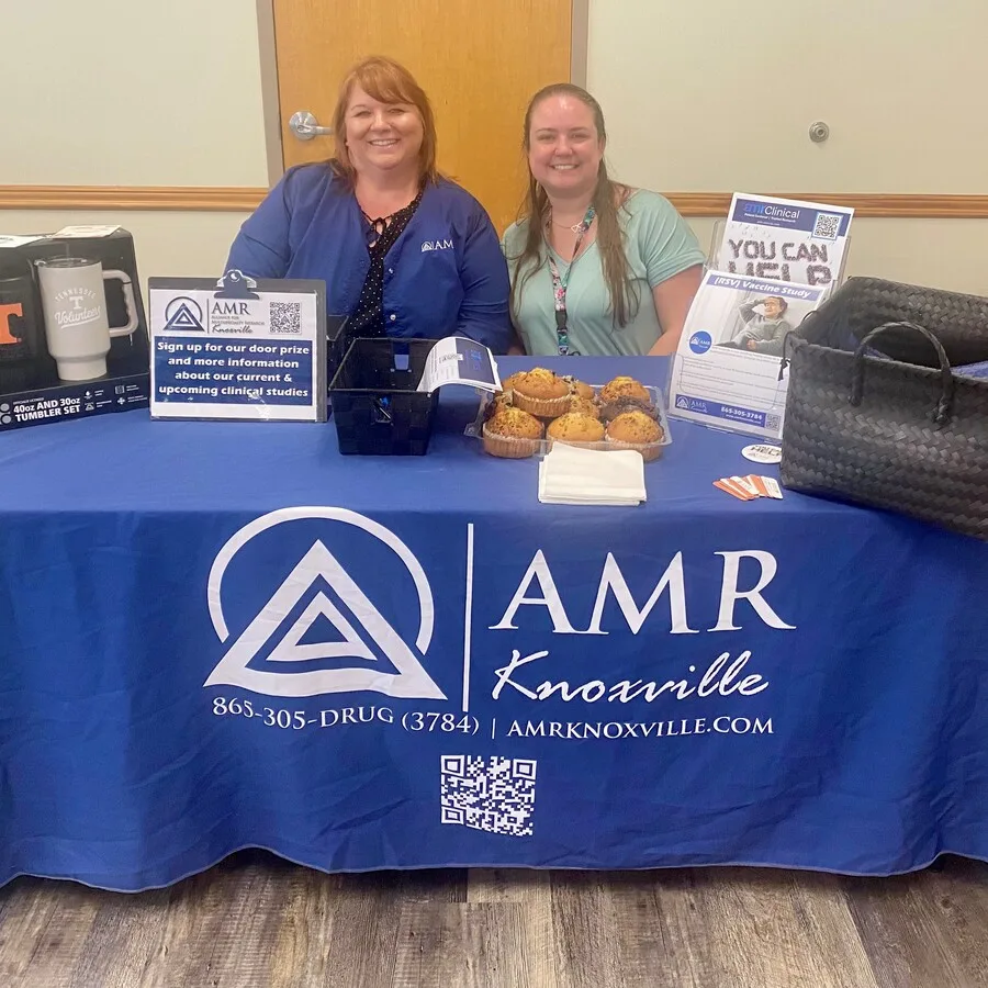 Two women sit behind an AMR Knoxville table at a senior job fair. The table is covered in a blue AMR-branded tablecloth and displays promotional materials, a sign-up form, muffins on a tray, and a basket for entries. A sign encourages attendees to register for door prizes and learn more about clinical studies. Promotional flyers and a “You Can Make a Difference” vaccine study poster are also visible.
