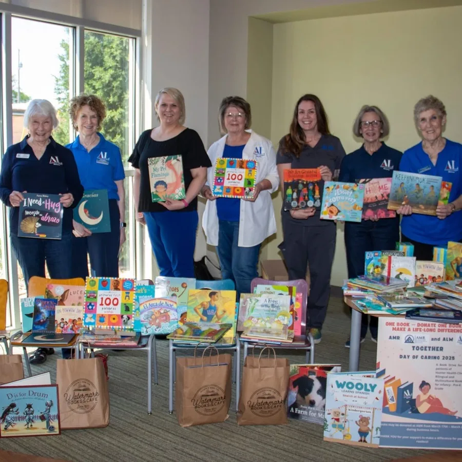 AMR team members pose indoors next to a long table filled with children’s books. Behind them, a “Day of Caring” sign is displayed. The table includes popular titles and decorations for the book donation drive.