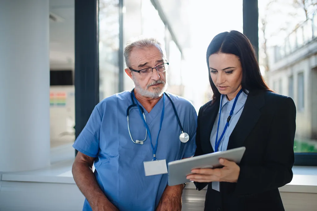 An older male doctor in blue scrubs with a stethoscope around his neck and a younger woman in business attire standing by a bright window, both looking at a tablet the woman is holding.