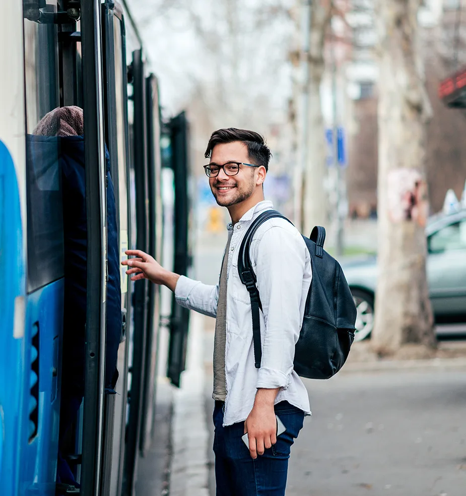 Smiling young man with glasses and a backpack boarding a blue city bus, looking back toward the camera on an urban street.