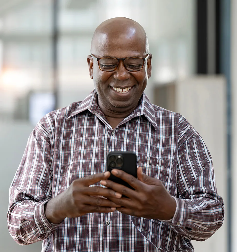 Man in a plaid shirt smiling while looking down at his smartphone in a bright indoor setting.