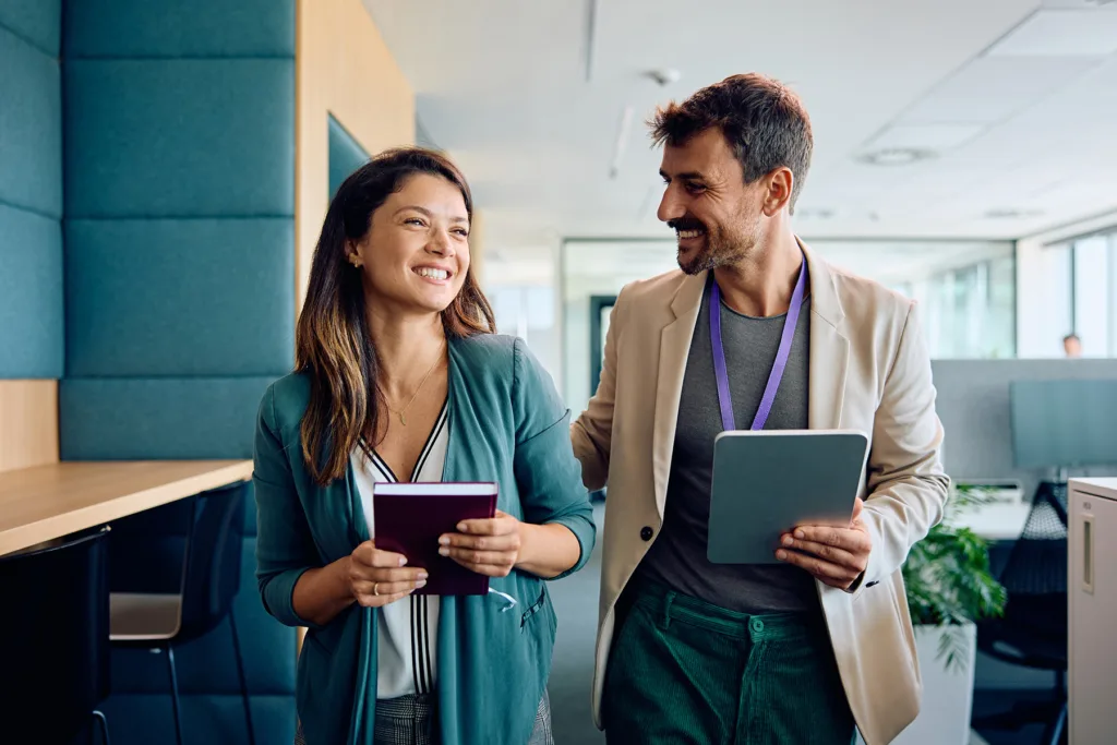 Two coworkers walking side by side in a modern office, smiling and engaged in conversation; a woman in a teal blazer holds a purple notebook while a bearded man in a beige jacket with a purple lanyard holds a tablet.