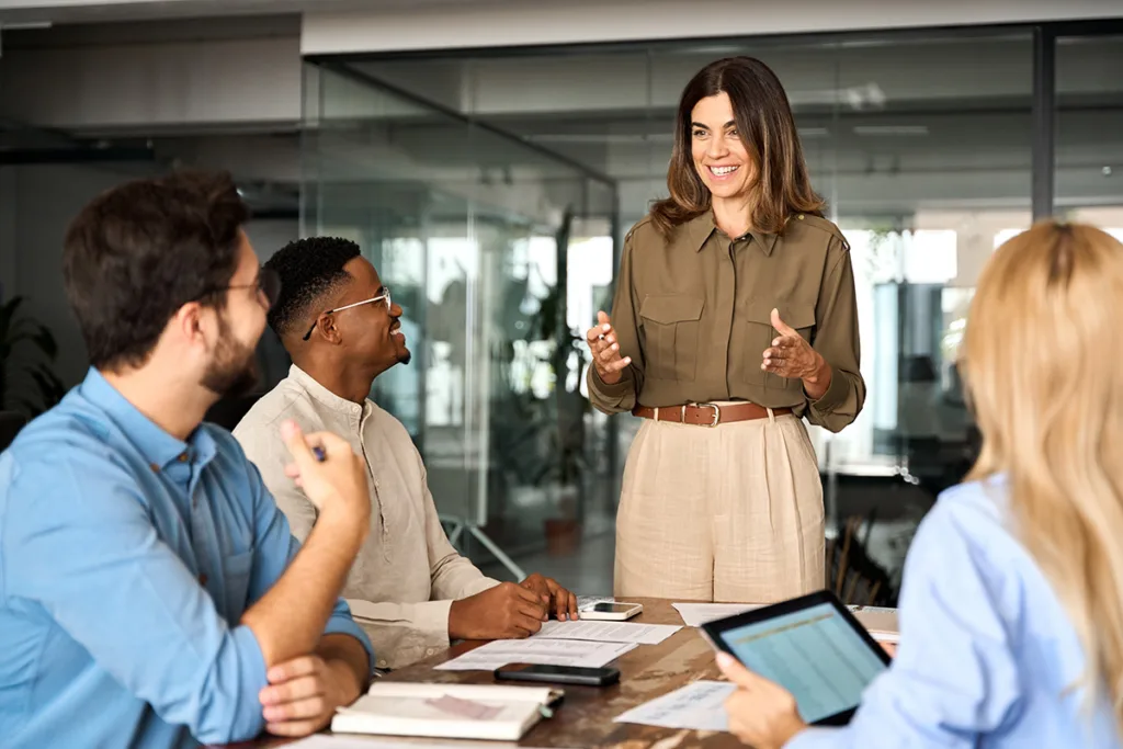 A smiling woman standing and speaking to three seated colleagues around a conference table; two men on the left are listening attentively and a blonde woman on the right holds a tablet, with papers and a smartphone on the table.