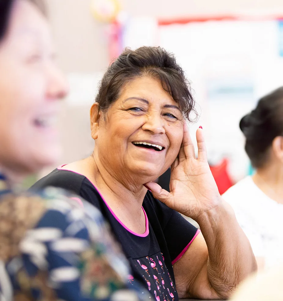 Older woman laughing with her hand to her ear during a group gathering, with other people softly blurred in the foreground.